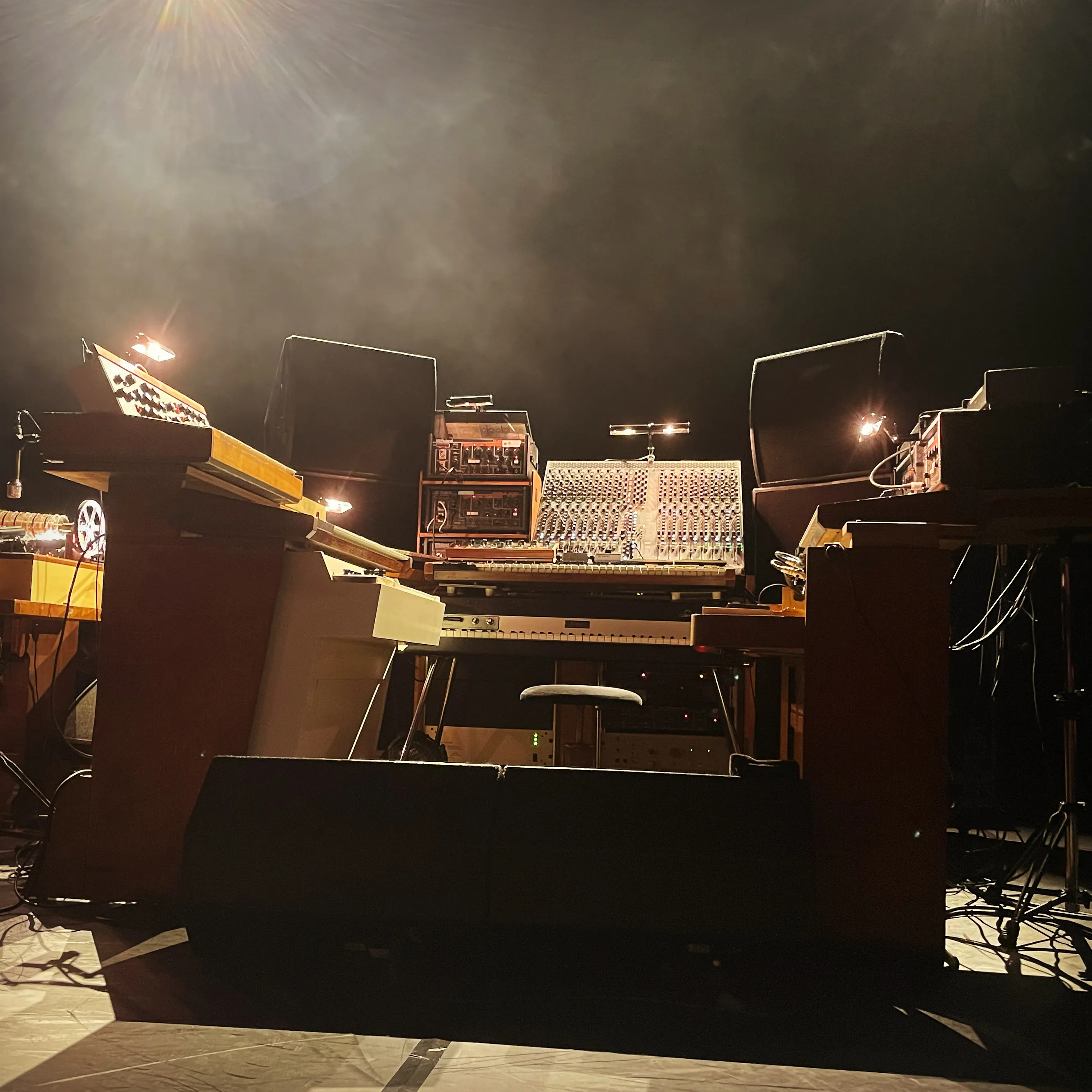 A stage of synthesisers, pianos and a mixing desk under warm light